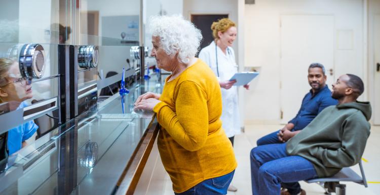 Senior woman talking with receptionist in waiting room in medical clinic. Two men talking with female doctor in the background.