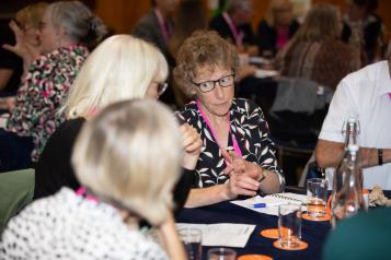 Jane Laughton is a middle-aged white woman. She is wearing glasses, a black and white shirt and has short, light-brown, curly hair. She is talk to other memebers of Healthwatch at a conference.