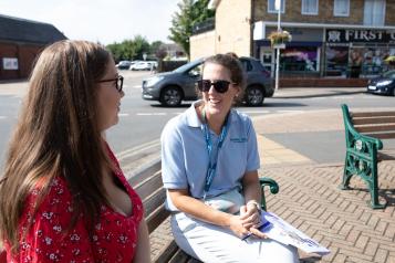 Two women are sitting on a bench outside on a sunny day. They are talking to each other. One is a Healthwatch employee wearing a blue shirt and sunglasses and facing towards the camera. The other is facing away and wearing a red blouse.